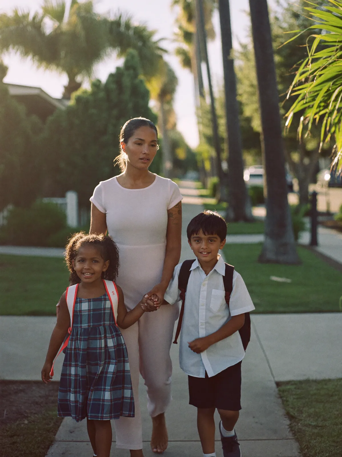 A mother walks her two young children along a palm-lined Tampa sidewalk in warm morning light, holding her daughter's hand